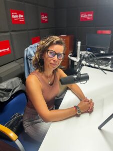 Studentka w studiu radiowym siedzi przy mikrofonie, przy stole ; ma krótkie włosy i okulary ; w tle banery promocyjne: Radio Elka i miedziowe.pl