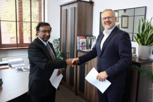 Rektor PANS w Głogowie, dr Jarosław Hermaszewski oraz Prezes American Medical Academy – John K. Eapen w gabinecie Rektora. Ubrani w garnitury, podają sobie prawe dłonie, a w drugiej ręce trzymają podpisany dokument: list intencyjny o współpracy pomiędzy instytucjami.
