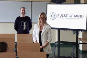 Mgr Anna Akielaszek-Witczak – Uczelniany Koordynator Programu Erasmus+.i przedstawiciel Pulse of Mind: Mindaugas Latisenka podczas spotkania z pracownikami, w auli, w tle widać telewizor z prezentacją.