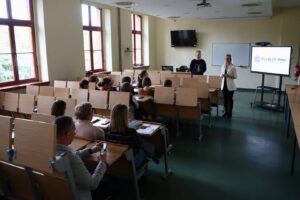 Mgr Anna Akielaszek-Witczak – Uczelniany Koordynator Programu Erasmus+.i przedstawiciel Pulse of Mind: Mindaugas Latisenka podczas spotkania z pracownikami, w auli, w tle widać telewizor z prezentacją.
