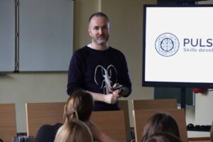 Przedstawiciel Pulse of Mind: Mindaugas Latisenka podczas spotkania z pracownikami, w auli, w tle widać telewizor z prezentacją.