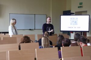 Mgr Anna Akielaszek-Witczak – Uczelniany Koordynator Programu Erasmus+.i przedstawiciel Pulse of Mind: Mindaugas Latisenka podczas spotkania z pracownikami, w auli, w tle widać telewizor z prezentacją.