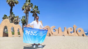 Uczelniany Koordynator Erasmus+ z PANS w Głogowie prezentuje flagę uczelni przy napisie „Malagueta” na plaży w Maladze podczas mobilności szkoleniowej.