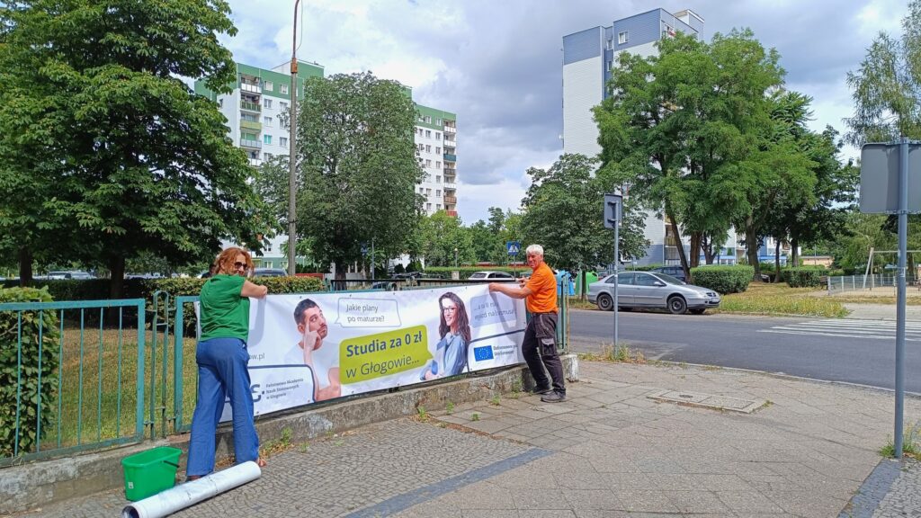 Pracownicy uczelni (kobieta i mężczyzna) rozwieszają baner reklamowy na płocie głogowskiej szkoły średniej. Baner przedstawia: zdjęcie studenta i studentki oraz ich rozmowa w formie dymków: Jakie plany po maturze? Odpowiedź: Studia za 0 zł w Głogowie, a na II roku wyjazd na Erasmusa. W lewym dolnym rogu Logotyp PANS w Głogowie, w prawym dolnym rogu Logotyp z flagą UE z informacją o dofinansowaniu przez Unię Europejską.