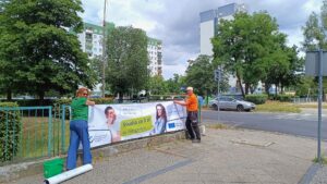 Pracownicy uczelni (kobieta i mężczyzna) rozwieszają baner reklamowy na płocie głogowskiej szkoły średniej. Baner przedstawia: zdjęcie studenta i studentki oraz ich rozmowa w formie dymków: Jakie plany po maturze? Odpowiedź: Studia za 0 zł w Głogowie, a na II roku wyjazd na Erasmusa. W lewym dolnym rogu Logotyp PANS w Głogowie, w prawym dolnym rogu Logotyp z flagą UE z informacją o dofinansowaniu przez Unię Europejską.