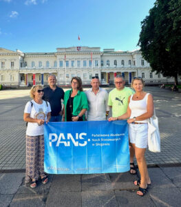 Mobilność szkoleniowa pracowników w ramach programu Erasmus+ do Wilna ; sześć osób (trzy kobiety i trzech mężczyzn) z niebieską flagą z logotypem PANS w Głogowie oraz Erasmus+ pozują na tle zabytkowego budynku w Wilnie