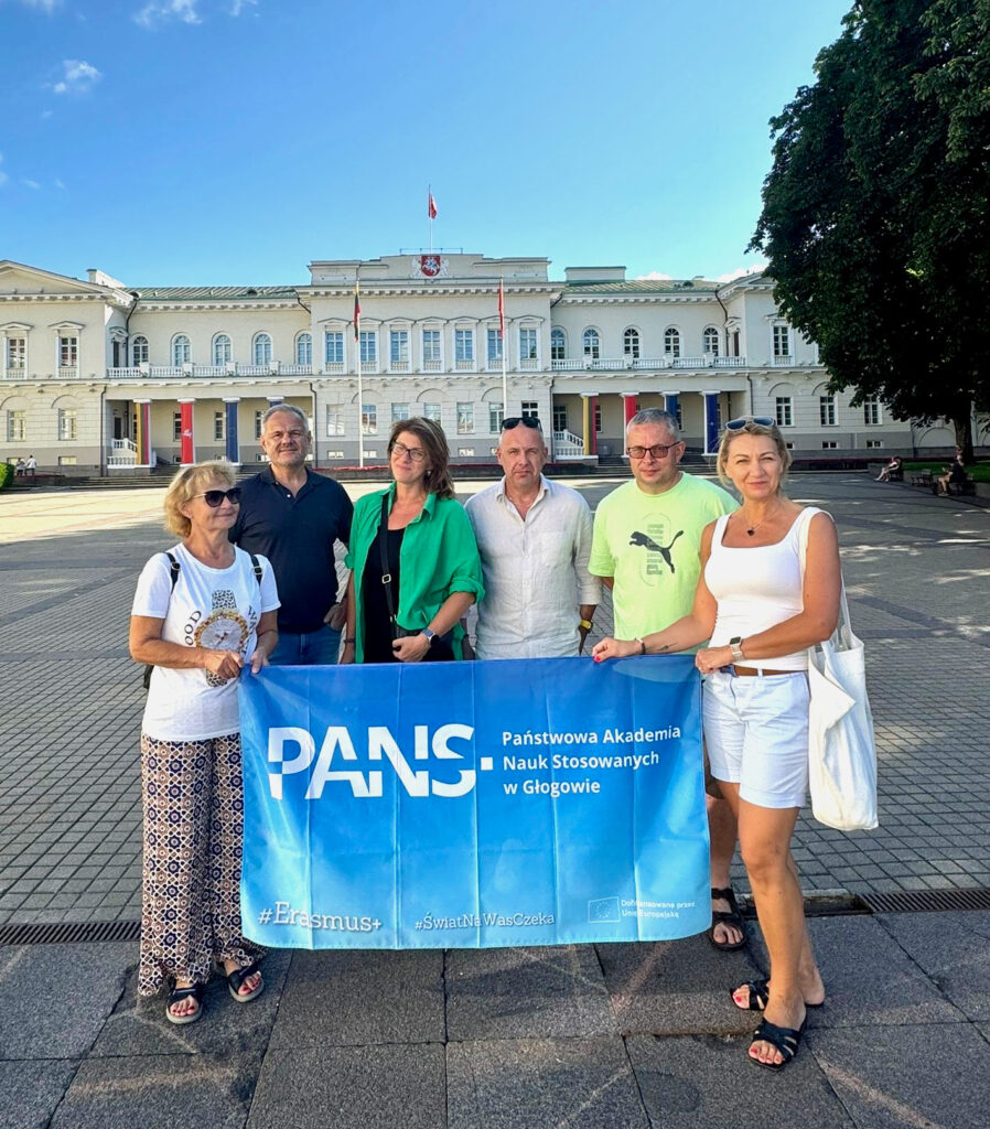 Mobilność szkoleniowa pracowników w ramach programu Erasmus+ do Wilna ; sześć osób (trzy kobiety i trzech mężczyzn) z niebieską flagą z logotypem PANS w Głogowie oraz Erasmus+ pozują na tle zabytkowego budynku w Wilnie